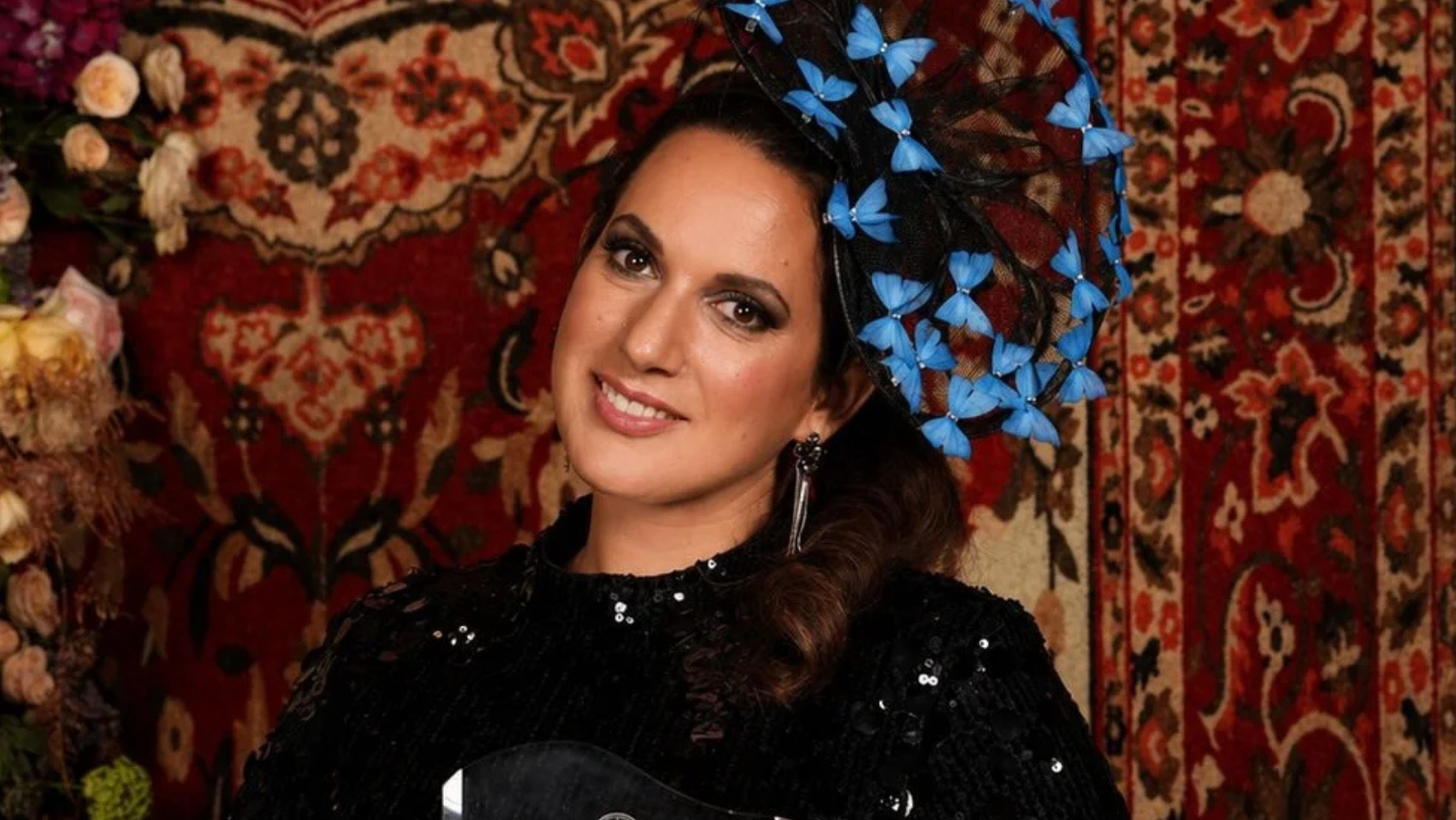 Award winner poses elegantly in a sequined outfit and decorative hat against a vibrant floral backdrop.