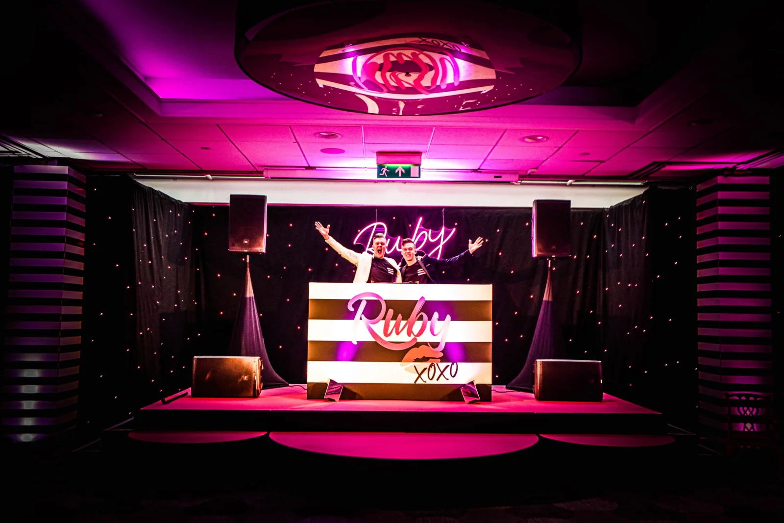 A vibrant DJ booth with pink lighting and a sign reading "Ruby" sets the festive atmosphere for a Bat Mitzvah celebration.